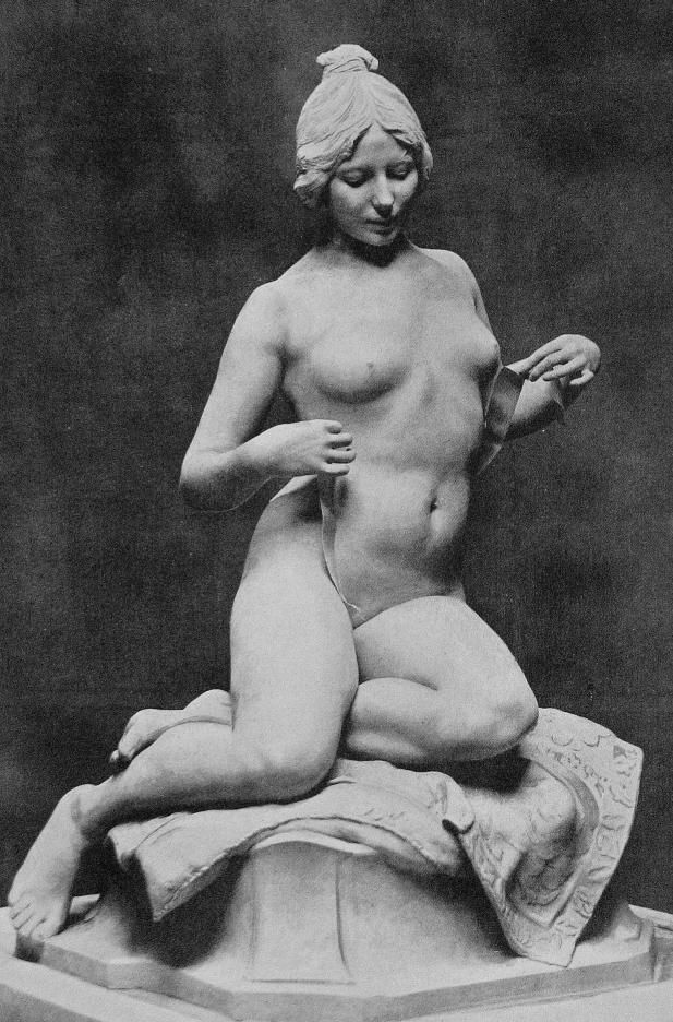 a black and white image shows a sculpture of a nude woman seated on a draped cloth placed on a pedestal. the woman has her hair styled in an updo and is looking down at a ribbon or strip of cloth that she is holding with both hands. the sculpture is classical in style and appears to be made of marble. the background is dark and blurred, making the sculpture the focal point of the image.