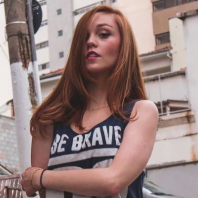 a young woman with long red hair is standing in an urban environment. she is wearing a black and white tank top that says "be brave" and has a black wristband on her left wrist. she is looking directly at the camera with a confident expression. the buildings in the background are blurred, creating a sense of depth. the lighting is natural and daytime, and the overall tone of the image is casual and stylish.