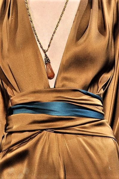 a woman is wearing a gold silk dress with a deep vneck. she is wearing a gold necklace with a gemstone pendant. the dress is belted with a dark blue sash. she is posed with the dress flowing around her.  the woman is looking to the left.