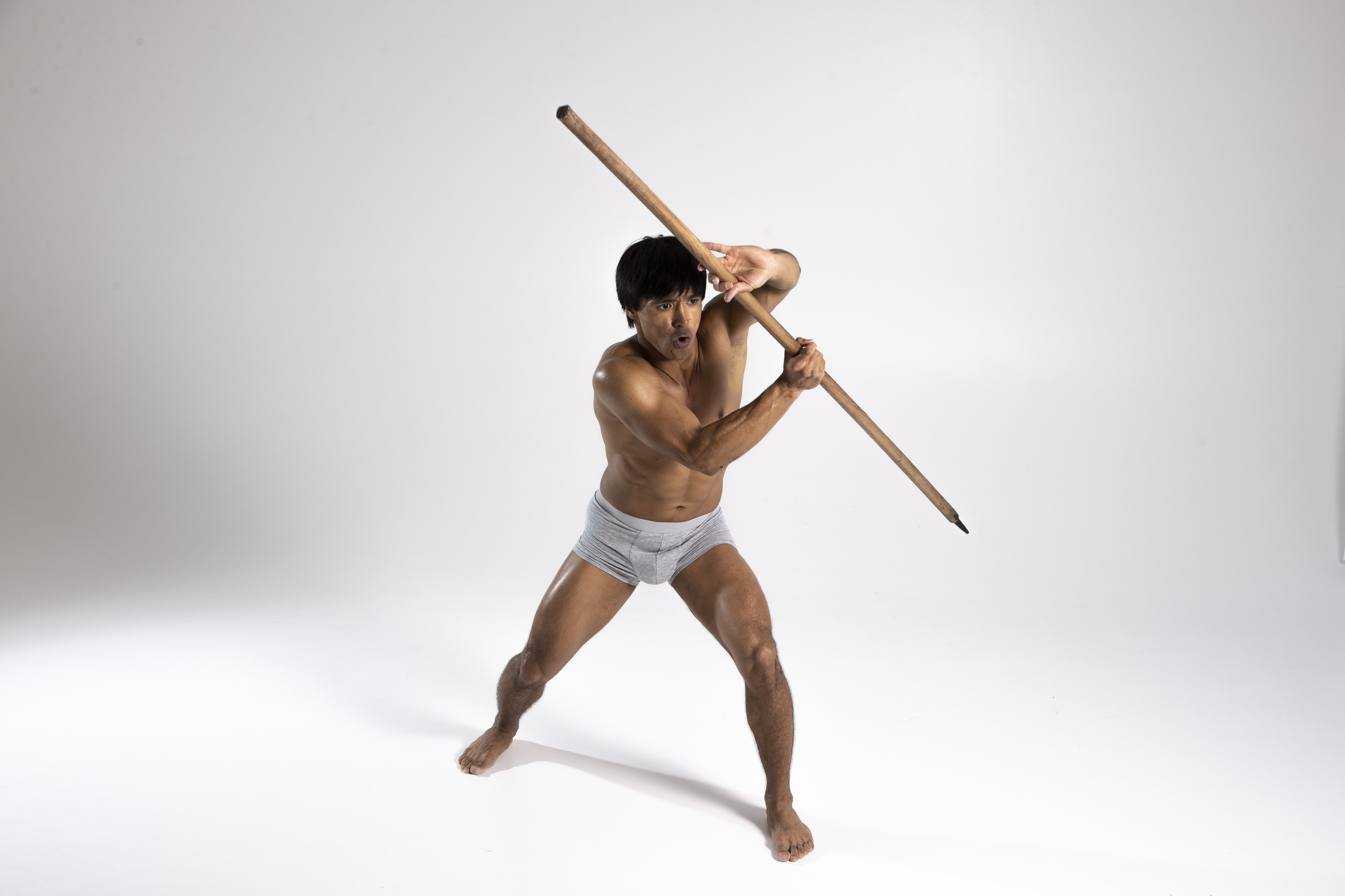 a shirtless, muscular man wearing gray underwear is holding a long, brown stick in a defensive stance. he is barefoot and standing on a white background in a studio shot. his mouth is open and he has a determined expression on his face.