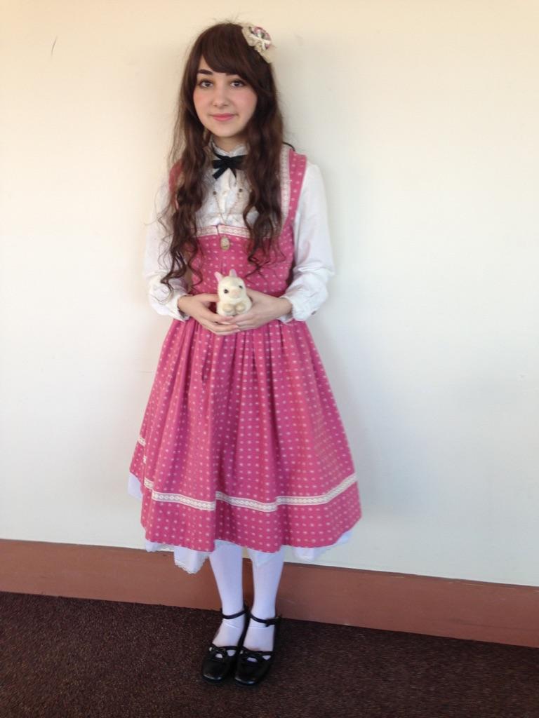 a fullbody shot of a girl wearing a pink polka dot dress with white lace trim.  she is wearing white socks and dark shoes and holding a small white stuffed rabbit. she has long brown hair and is wearing a small headband.  the photo is taken indoors against a light beige wall.  her dress is styled in a way reminiscent of vintage or lolita fashion.
