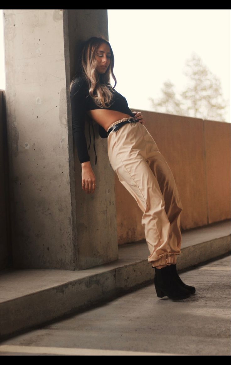 a stylish woman in a black shirt and beige pants is posing casually against a concrete wall. she is smiling, and the photo is taken in an urban setting.  the sunlight is shining brightly, and the woman's hair is long and blonde. her outfit is trendy and fashionable. she is wearing black boots.