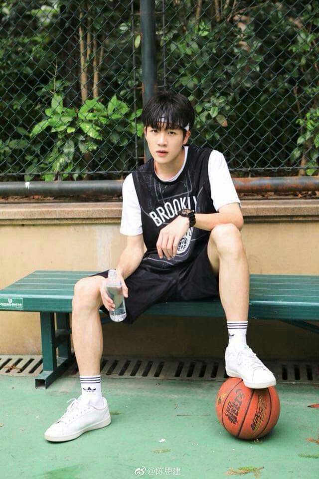 a young man in athletic wear is taking a break from playing basketball. he is sitting on a bench on an outdoor court, holding a water bottle. he has a basketball at his feet. the image is taken from an eyelevel perspective and the lighting is natural.