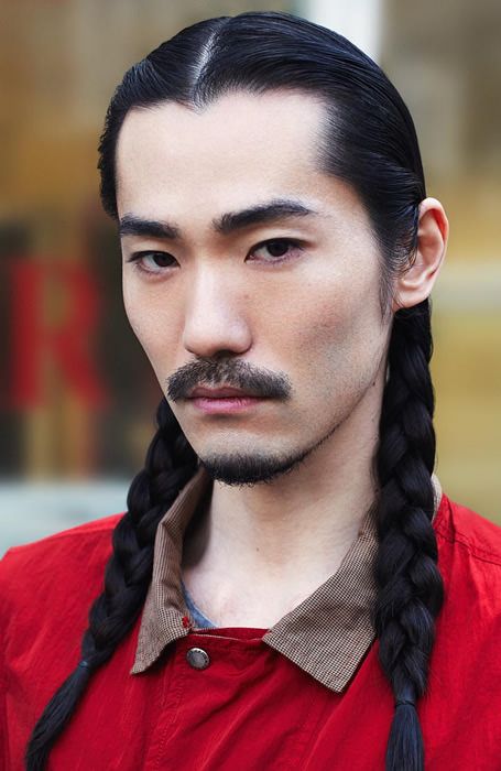 a closeup portrait of an asian man with black hair styled in two braids, a mustache, and wearing a red shirt with a buttoned collar. he has a serious expression.