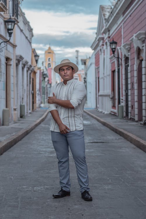 a fullbody shot of a man standing in a narrow alleyway between old colonial buildings. he is wearing a beige fedora hat, a light beige longsleeved shirt with thin vertical stripes, grey trousers, and black shoes.  the man is looking towards the camera with a serious yet confident expression. the architecture of the street suggests a latin american or hispanic setting. the overall mood is somewhat casual yet elegant. the image is welllit, presumably taken during the daytime.