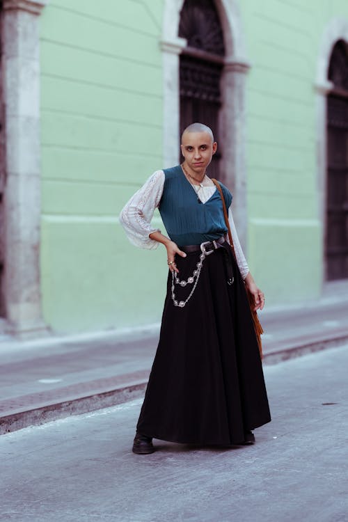 a bald woman stands on a city street in front of a light green building. she is wearing a teal vest over a white lace top, a long black maxi skirt, and a silver chain belt. she is carrying a brown fringed bag over her shoulder. she is looking directly at the camera.