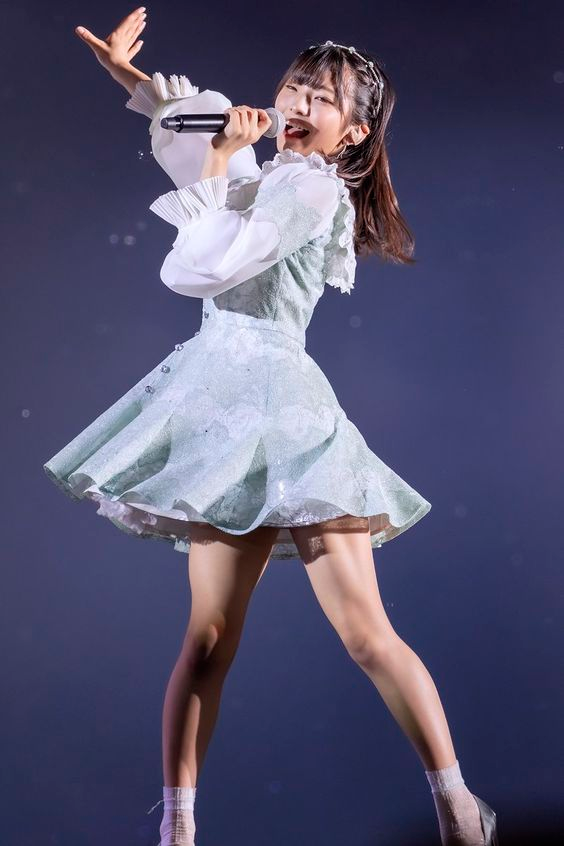 a young woman in a green and white dress is performing on stage. she's holding a microphone in her right hand and appears to be singing. the stage lighting casts a blue hue on the background, creating a contrast with her brightly lit figure.