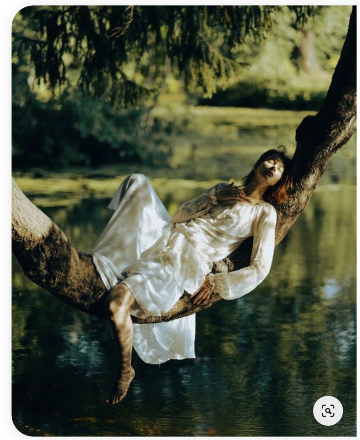 a woman in a white dress is lying on a tree branch that extends over a lake. she appears to be resting peacefully with her eyes closed. the scene is filled with natural light, reflecting on the water and illuminating the surrounding foliage.