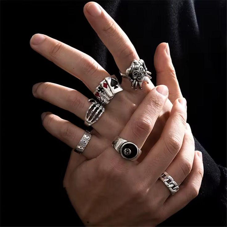 a closeup shot of a person's hand adorned with multiple silver rings. the rings have various designs including a skull, playing cards with an ace and spade, an eightball, a chain pattern, and a skeleton hand. the hand is set against a dark background. the image suggests a fashion or jewelry advertisement.