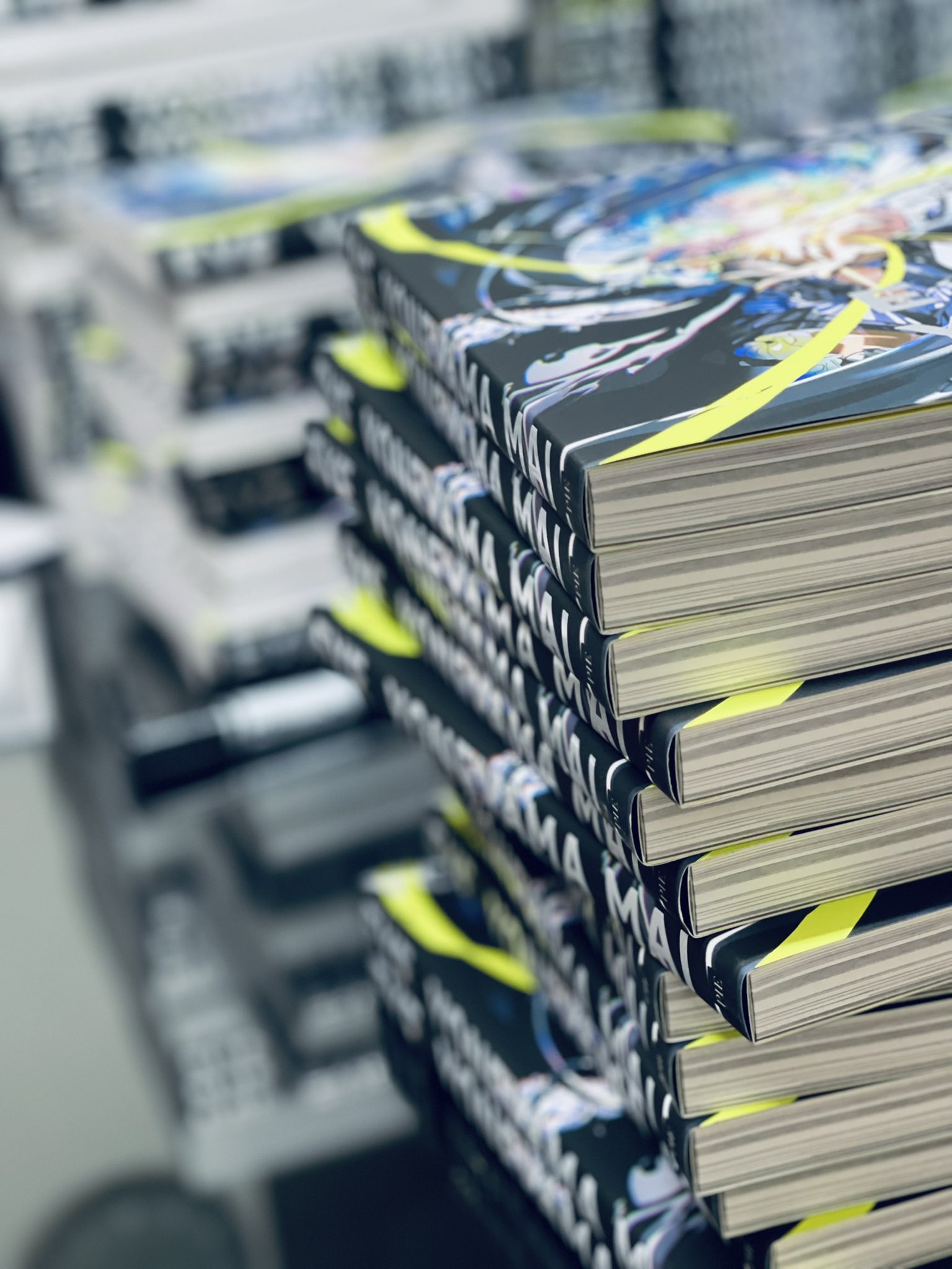 a stack of art books on a shelf with a shallow depth of field.  the book on top has a colorful, abstract design, and the spines of the books have repeating text that reads "mai". the focus is on the books, with the background blurred.