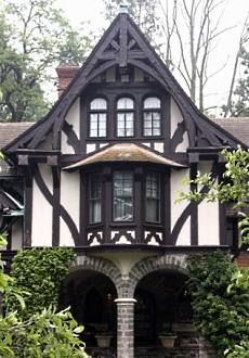 the image is of a tudor style house with dark brown timber framing and white stucco infill. the house has a steeply pitched gable roof with decorative woodwork. there are three arched windows on the second floor, and a bay window on the first floor. the entrance porch has two arches supported by brick pillars. the house is surrounded by trees and greenery.