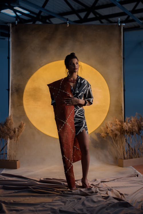 a fulllength shot of a stylish woman, seemingly a model, posing against a studio backdrop. she's wearing a chic, patterned dress with a darkcolored base and lighter, possibly white or beige, geometric designs.  she also wears a long, rustcolored textile draped over one arm and around her body. the dress appears to be a shortlength design, and the fabric has an almost ethnic or bohemian feel. she is accessorized with jewelry, including earrings and a necklace. the backdrop features a large, golden circle reminiscent of a sun, providing a warm, evocative ambiance.  the overall scene evokes themes of african or global style.  neutralcolored dried plants are visible in the background, adding to the styling. the floor is covered with a soft, lightcolored fabric. the setting suggests a professional fashion photoshoot.