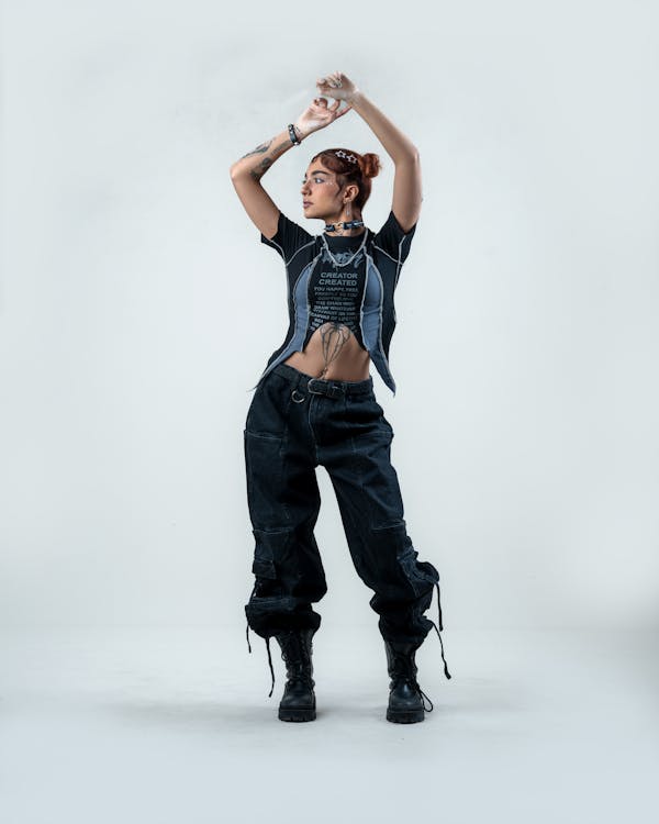 the image shows a young woman with red hair and tattoos posing against a white studio background. she's wearing a darkcolored, distressed denim top and cargo pants, black boots, a choker necklace and several rings and bracelets. her overall style is edgy and alternative, with a grunge influence.  her pose is confident and expressive, with her arms raised in a dynamic movement. the lighting and composition focus on her and her clothing, highlighting her style.