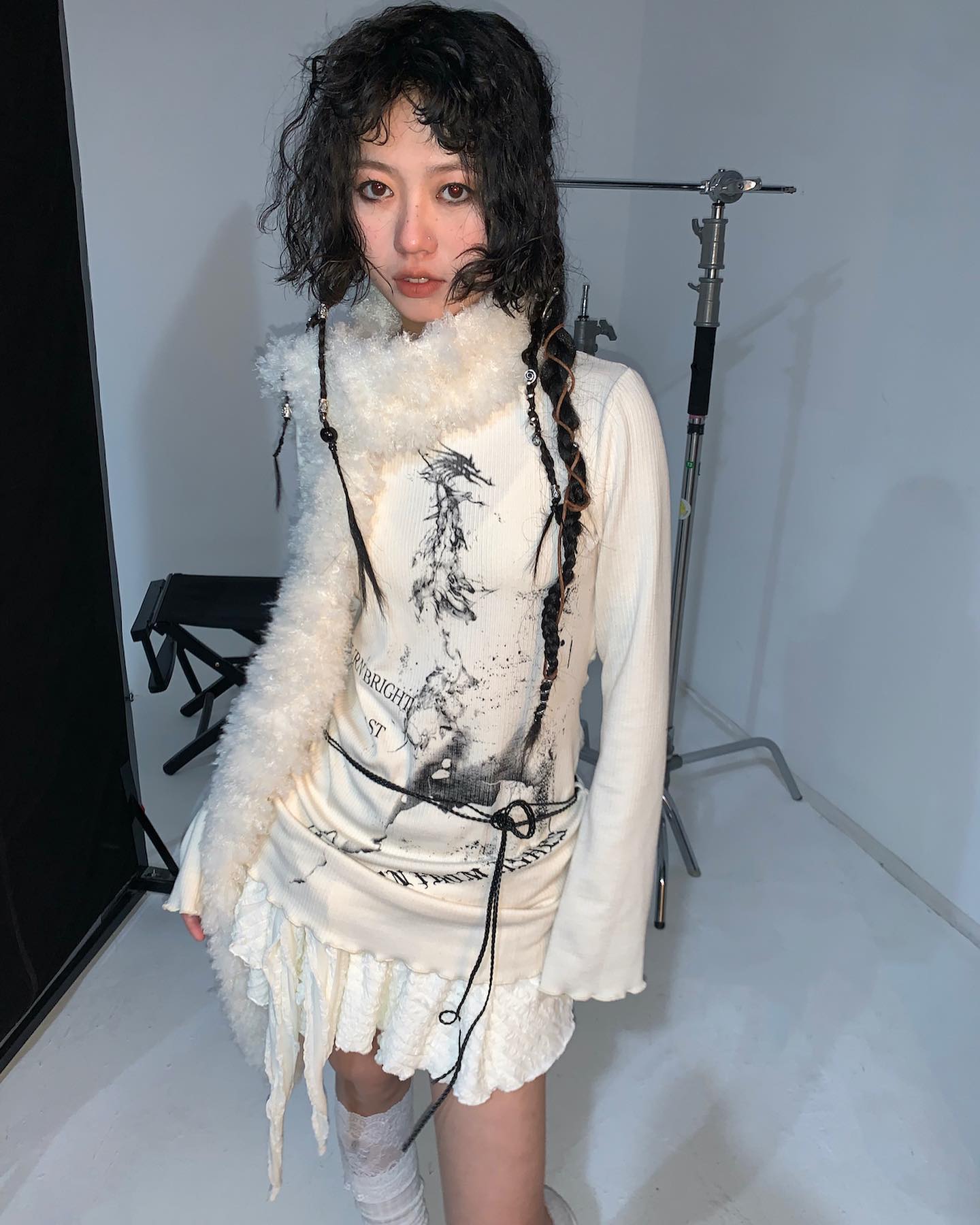 a young asian woman with dark curly hair models a unique white dress.  the dress features long sleeves, a ruffled hem, and a layered design.  a fluffy white collar and a black belt with a cord accent the garment. the dress has a graphic print, suggesting a grunge or gothicinspired style.  she is posing in a studio setting, with photography equipment visible in the background. the overall aesthetic is alternative and indie.