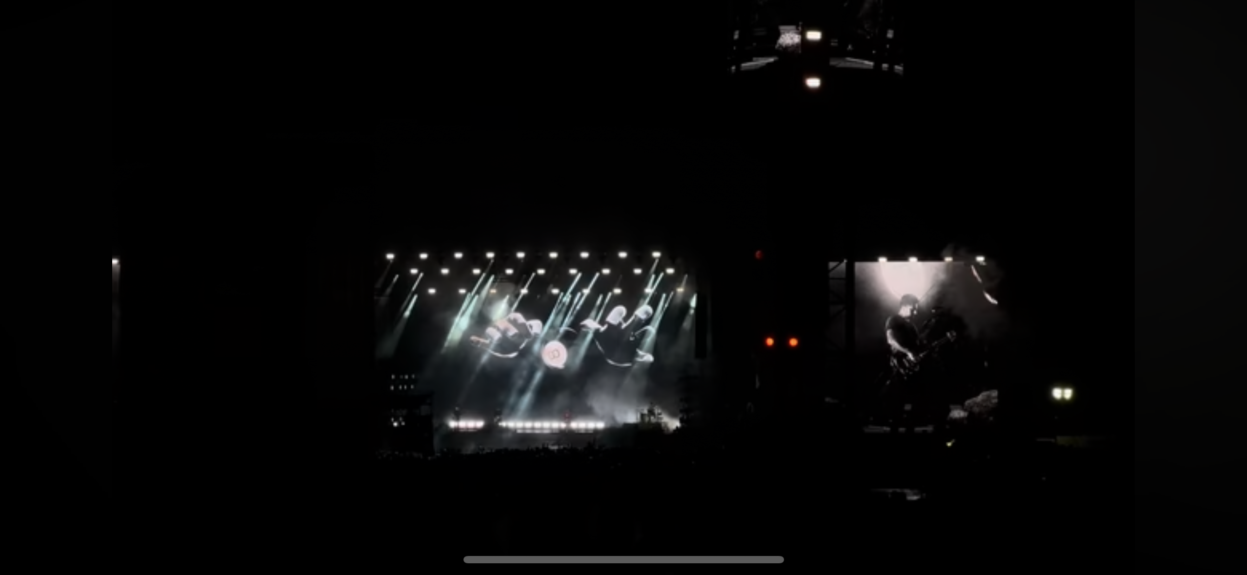 a dark concert venue with a large stage and a large screen playing a video of the headlining band performing.  a crowd of silhouettes stand before the stage,  enjoying the music and the energy of the show.  the stage is lit up with many white spotlights with light beams extending across the audience.  the image is captured from behind the crowd, giving a view of the show from a concertgoer's perspective.