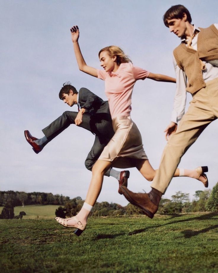 a young woman in a pink shirt and tan skirt runs and jumps in a field with two young men. the woman's hair is blonde. the men wear suits and dress pants. they all appear to be having fun and laughing. the sky is a clear blue and the grass is green.