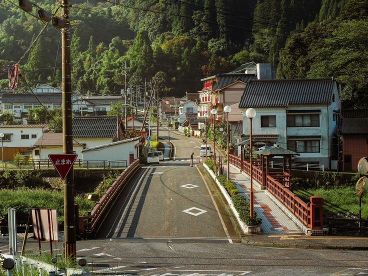 this image depicts a road in a japanese town. the road leads uphill, lined with houses and street lamps. a pedestrian walkway runs alongside the road on a bridge with red railings. a stop sign is visible on the left side of the road. trees cover the hillside in the background. several electrical or utility poles are also visible. a person is walking in the middle of the road.