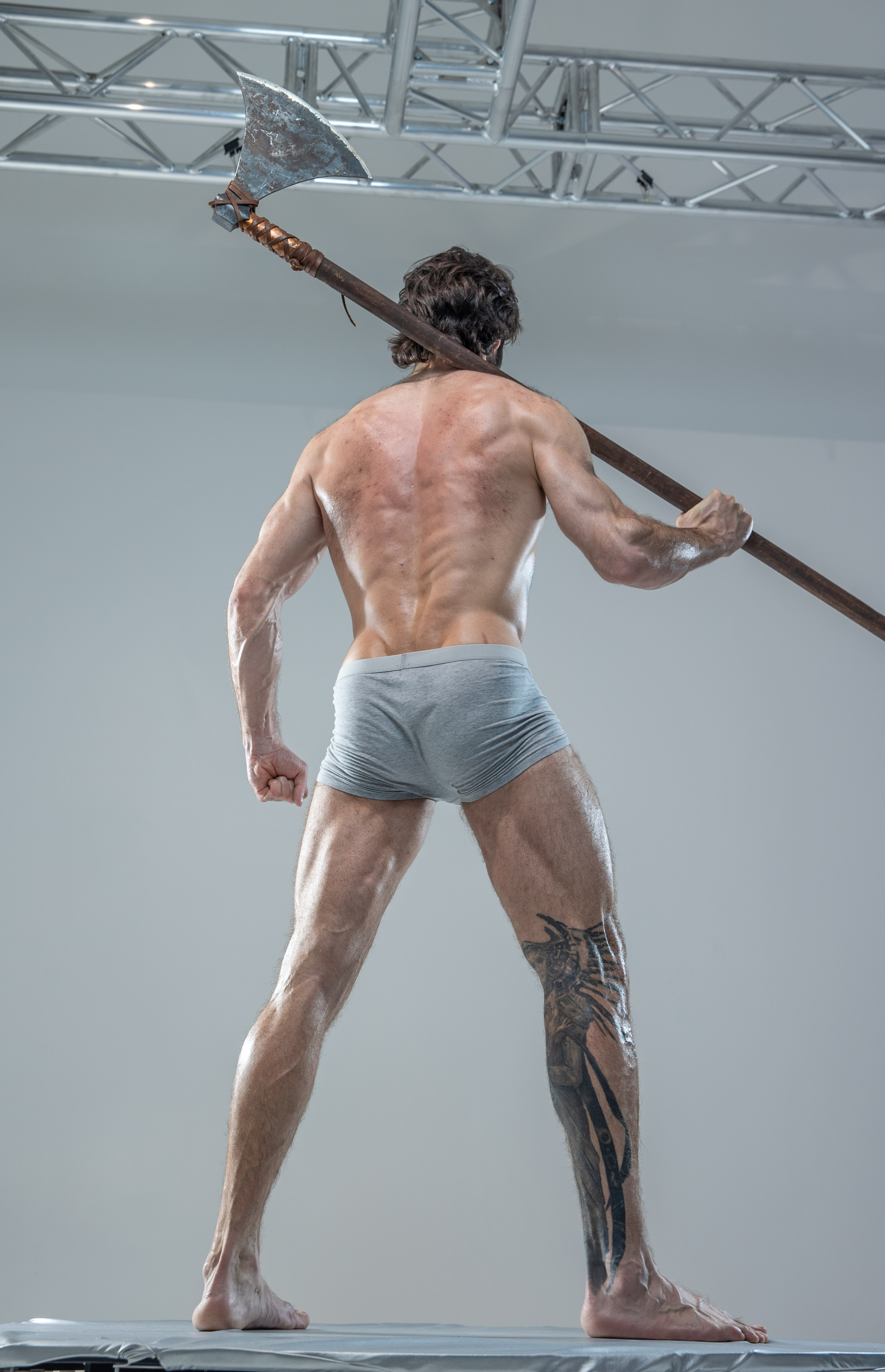 a muscular man, shot from a low angle and from behind, stands holding a large axe over his shoulder. he is shirtless, wearing only grey underwear, and has a tattoo of a dragon on his left calf. he is standing on a platform in a studio setting with bright, artificial lighting.