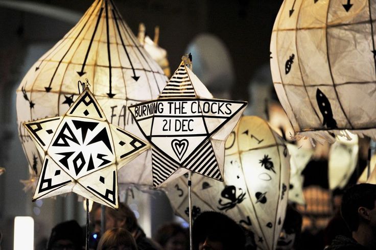 a crowd of people holding lanterns, including starshaped lanterns with "burning the clocks", "21 dec", "2015", and other designs during a night parade or festival.