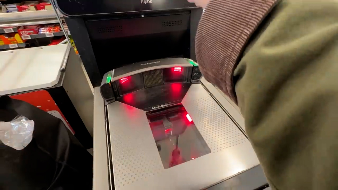 a person is using a selfcheckout kiosk at a grocery store. the image shows the scanner with red lights activated as a hand moves to scan an item. the perspective is from the shopper's point of view.