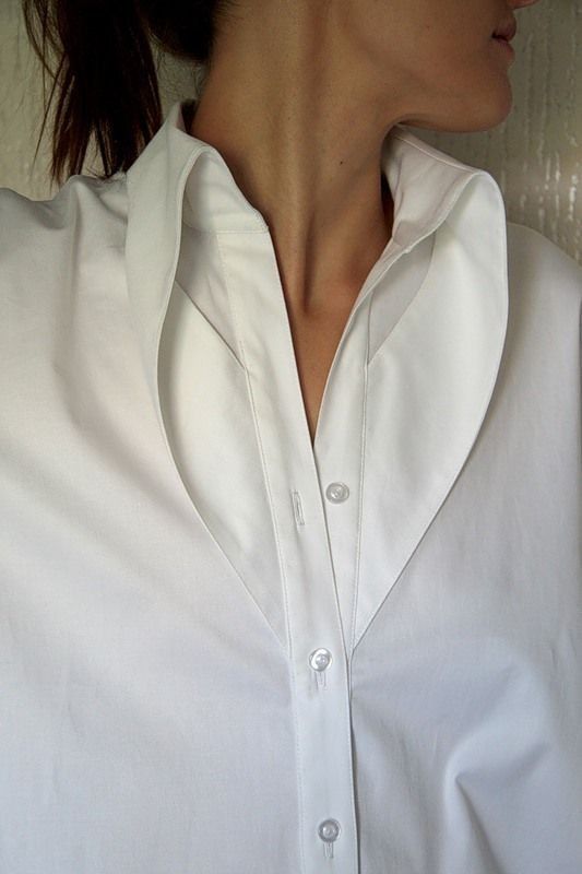 a closeup shot of a woman wearing a white collared shirt. the shirt is partially buttoned, and the collar is wide and open. the focus is on the upper part of the body, highlighting the neck, collarbone, and the shirt's design. the skin tone and the white fabric create a clean and simple aesthetic.