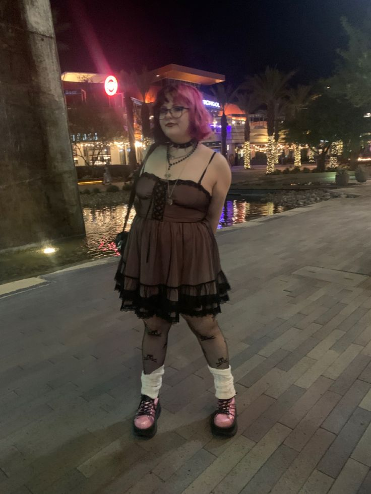 a woman with pink hair, a choker, and necklaces is standing outside on a brick path near a shopping center at night. she is wearing a black and brown dress, patterned tights, leg warmers, and pink platform shoes. there are lights, water, and trees in the background.