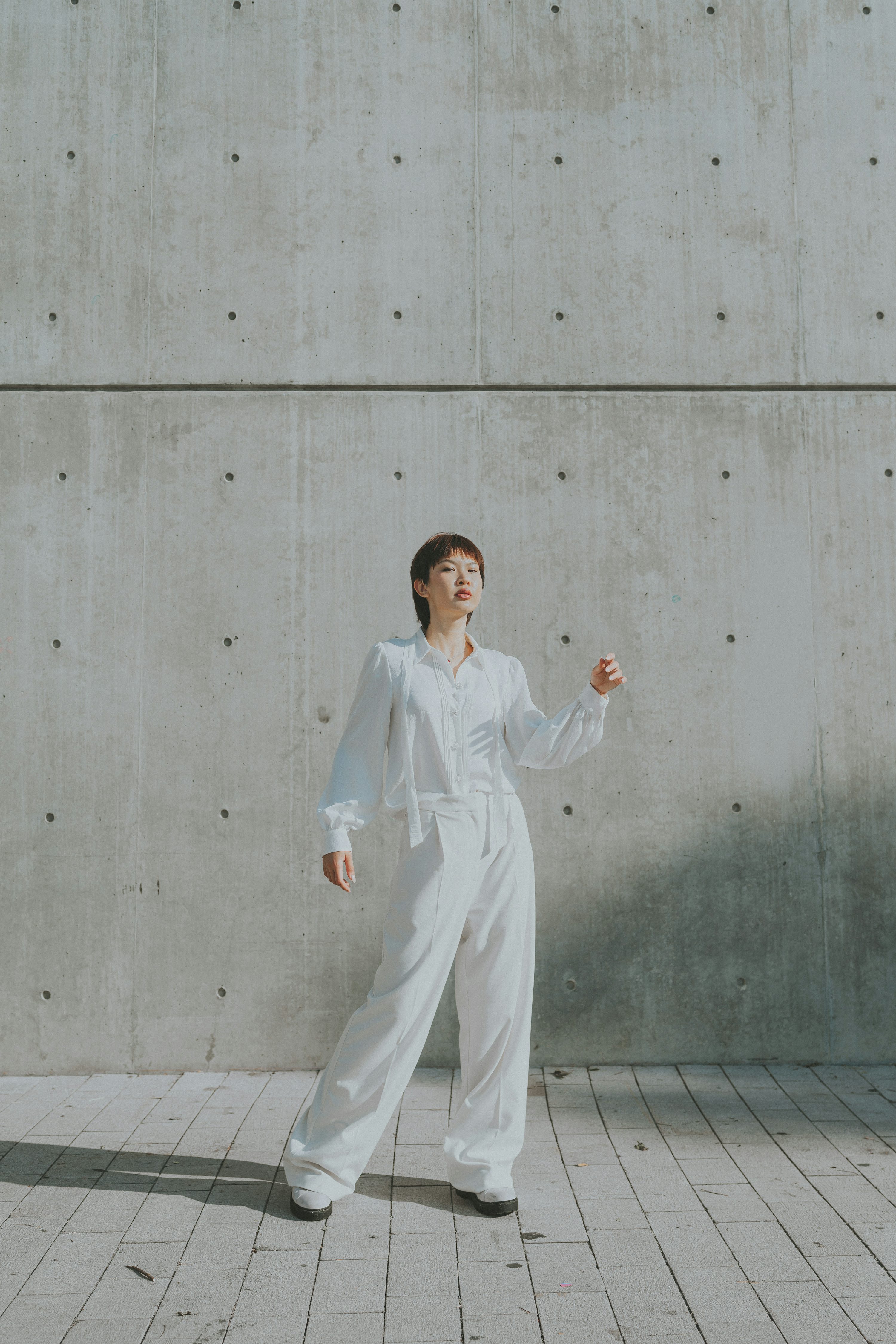 a woman stands in front of a concrete wall, wearing an allwhite outfit consisting of a shirt and pants. she has short hair and is looking up. the scene is lit by sunlight.