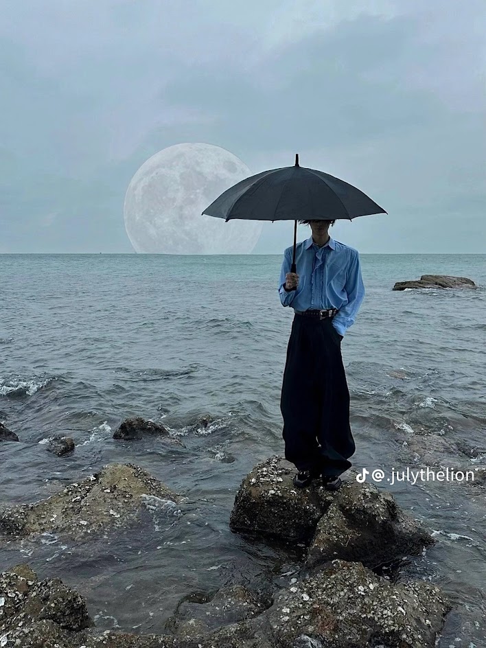 a man stands on a rock by the ocean holding a black umbrella as the full moon looms behind him.  the man is dressed in a blue shirt and black pants. the sky is overcast and the water is a dark blue. the man is looking off to the side and there are rocks and pebbles on the beach around him.  the scene has a cool and moody vibe.