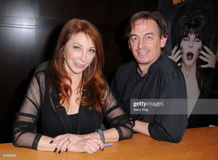 a woman with red hair and a black dress is sitting at a table with a man with black hair and a black shirt. they are both smiling. the woman is signing an autograph for a fan. they appear to be at an event or convention.