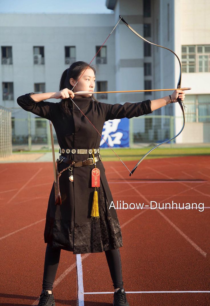 the image shows a young asian woman, possibly of chinese descent, dressed in a black traditionalstyle costume, aiming a recurve bow and arrow on an outdoor running track.  the bow and arrow appear to be of high quality. she is focused and poised, exhibiting a stance suggesting she's practicing or participating in archery.  the background is a modern building and a running track. a watermark "alibowdunhuang" is visible, possibly indicating the brand of the bow or the location.  the overall aesthetic blends traditional attire with a modern sporting setting.
