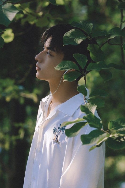 a young asian man is standing in a forest, surrounded by lush green foliage. he is wearing a white buttondown shirt with a tropicalthemed embroidery. he has earbuds in and appears to be listening to music or a podcast, with a serene and contemplative expression on his face. the soft, natural lighting filters through the leaves, creating a dappled effect on his face and clothing. the image evokes a sense of tranquility and connection with nature.