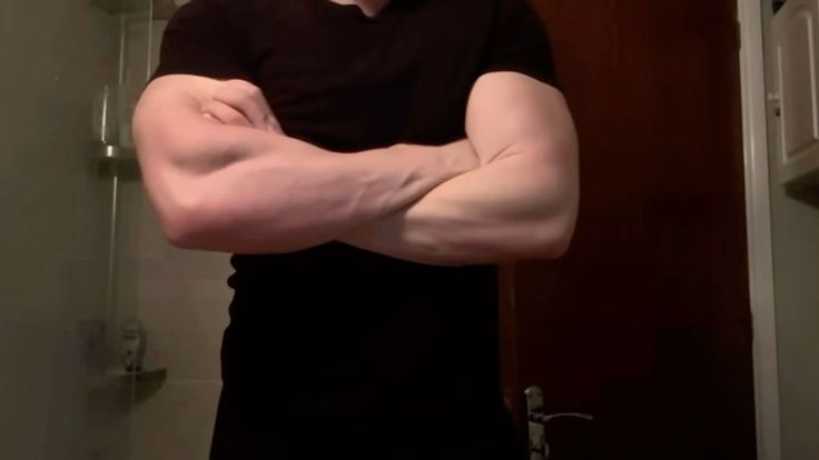 a closeup image of a muscular man with his arms crossed in front of him. he is wearing a black shirt and is standing in a bathroom. the man's biceps and triceps are clearly visible, showcasing his physique. the background is blurred, but a shower and part of a mirror are visible. the overall tone of the image suggests a selfportrait or a fitnessrelated photograph.