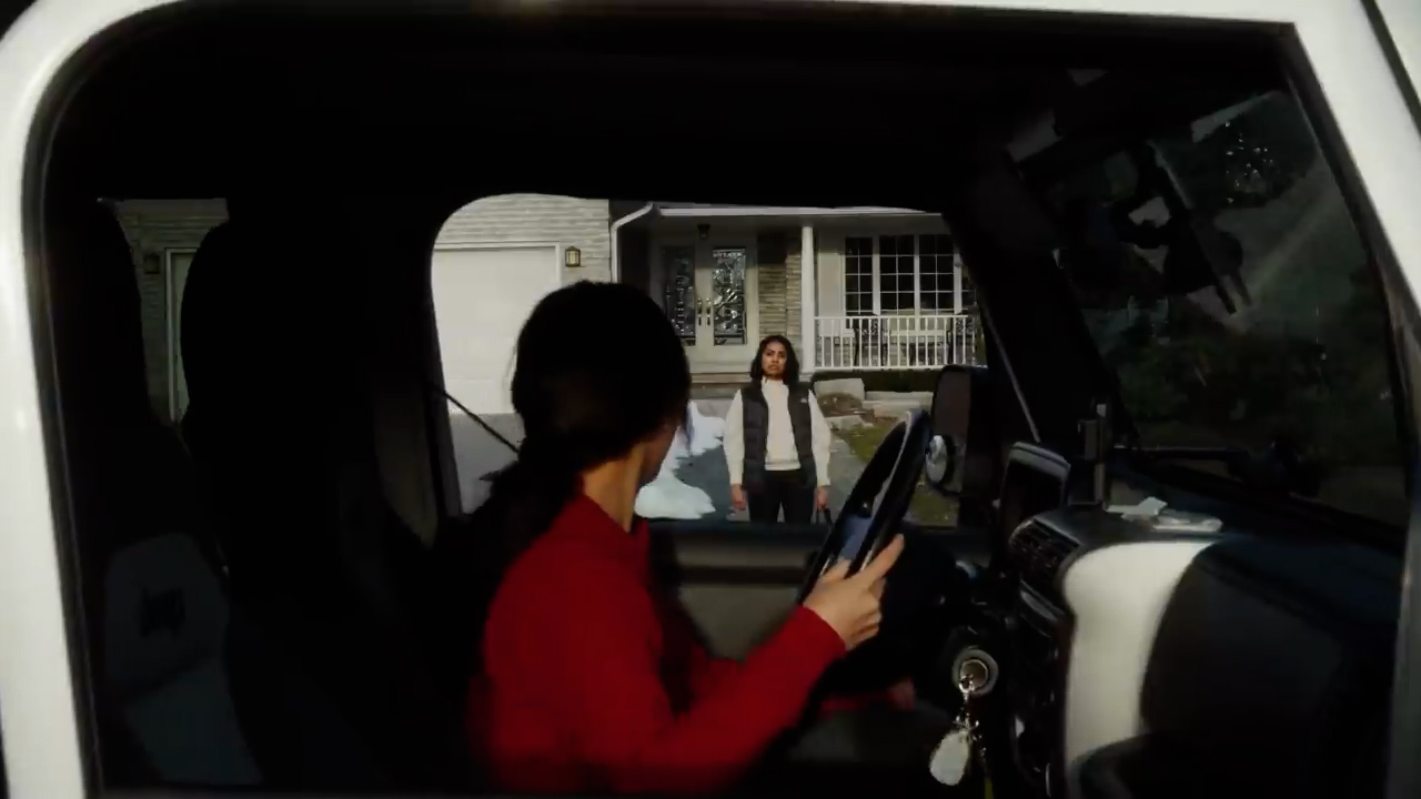 the image shows the interior of a car. the driver, a woman wearing a red shirt, is looking out the window. outside the car, another woman is standing on the sidewalk in front of a house. she is wearing a grey vest and holding a package. it appears the driver is about to pick her up. the photo is taken from the perspective of someone sitting in the passenger seat. the lighting is natural and overcast, giving the image a slightly muted feel.