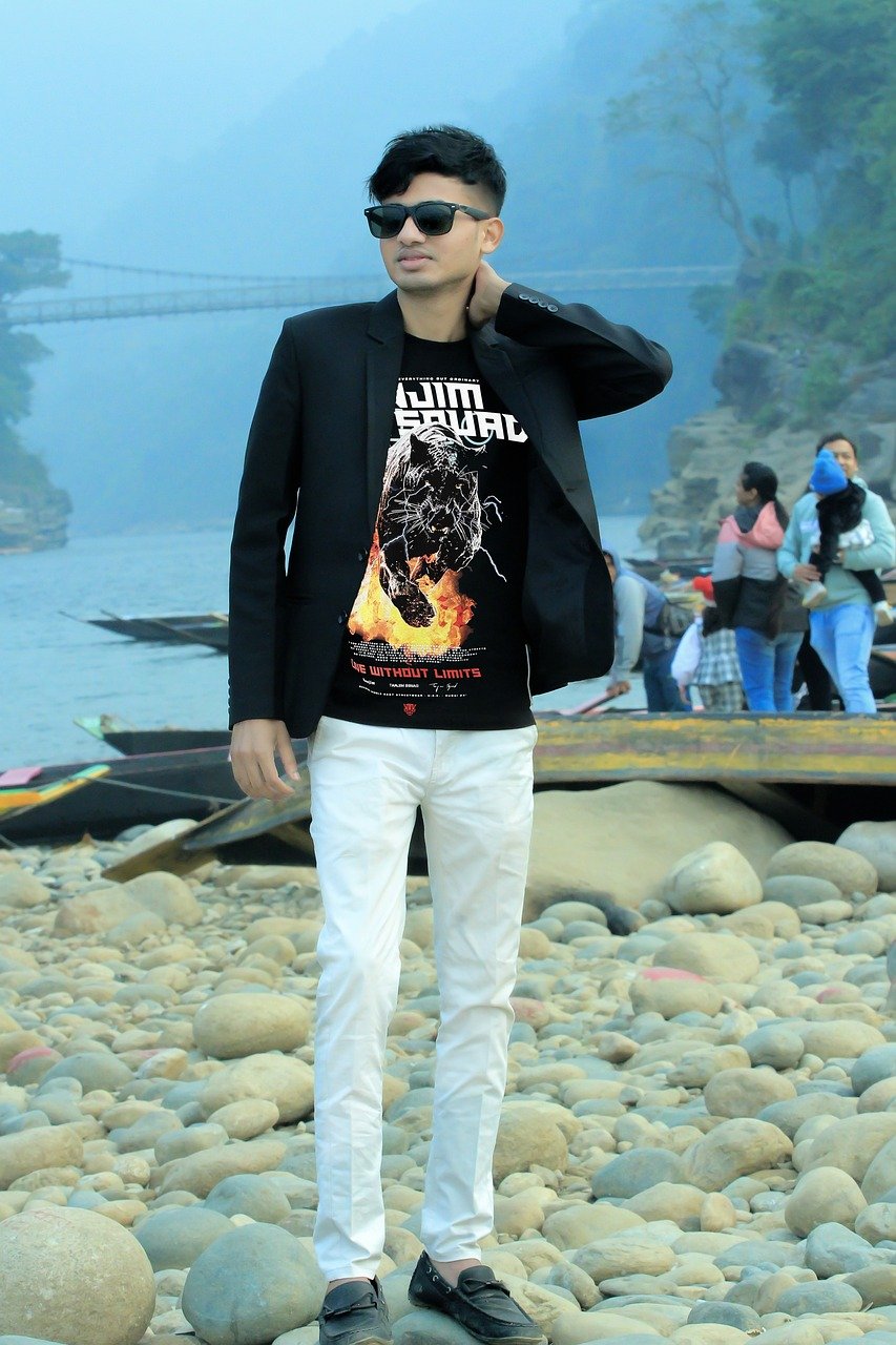 a young man in a black jacket and white pants stands on a rocky beach near a river. he is wearing sunglasses and looking towards the camera.  a bridge is visible in the background, along with mountains and a hazy sky.  other people are visible in the distance, but blurred, near boats.  the overall scene is casual and outdoors.