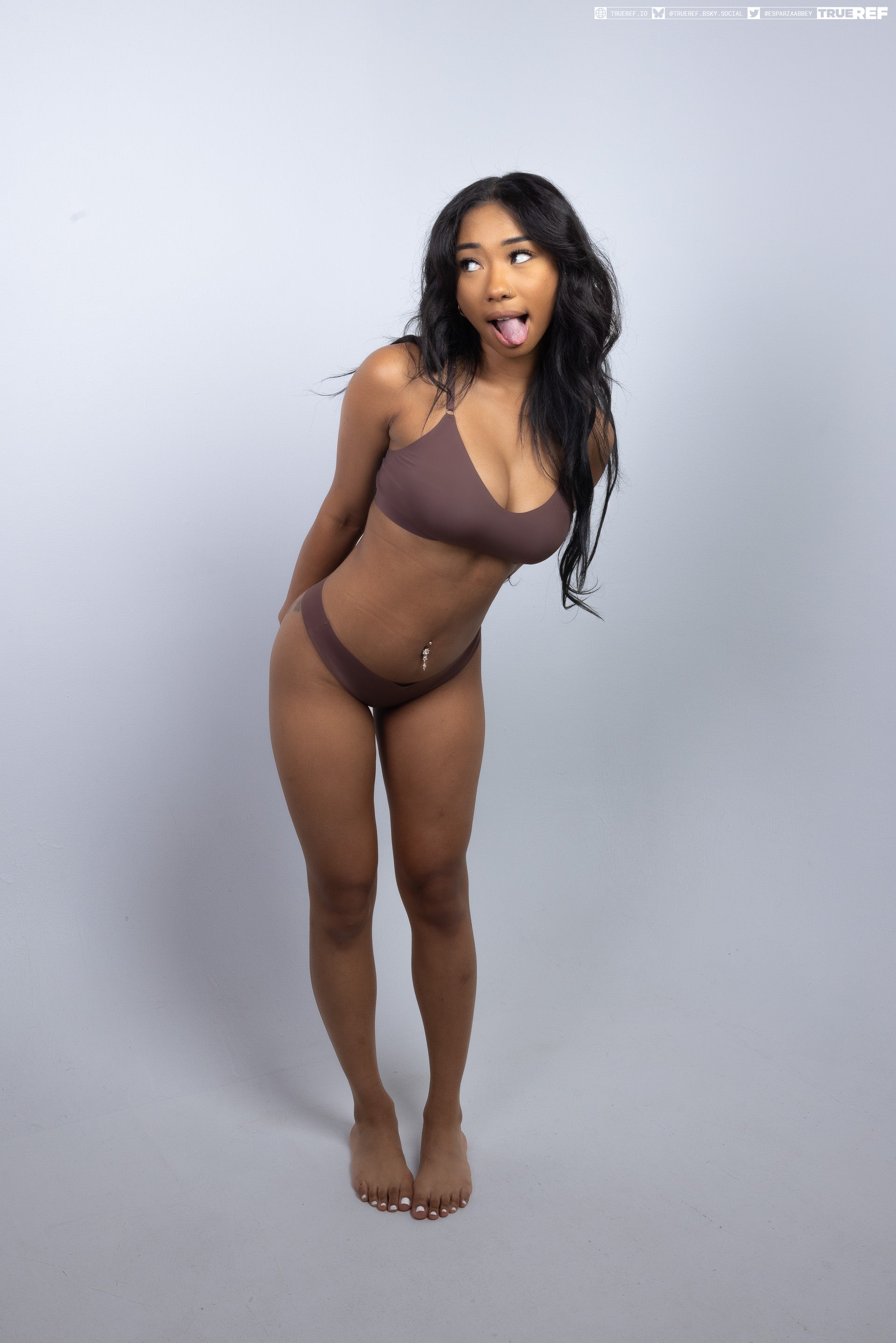 a fulllength studio shot of a slender, brownskinned woman wearing a brown lingerie set, standing barefoot with her tongue sticking out. she has a navel piercing and curly black hair. she is looking away from the camera with a playful expression.