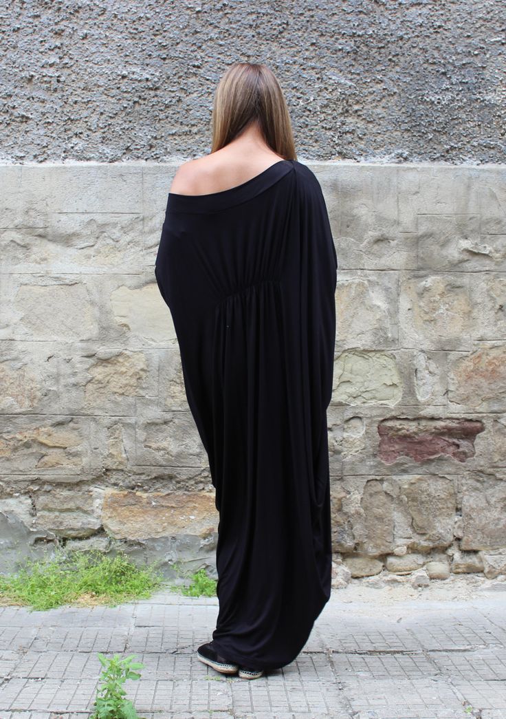 a woman stands with her back to the camera, wearing a long, black offshoulder dress, against a textured stone wall. the setting appears to be outdoors on a paved area.