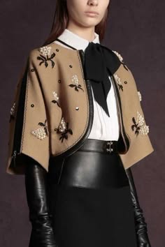 a woman is standing in front of a solid color backdrop. she is wearing a brown embroidered jacket with a black leather skirt and gloves. she is also wearing a white shirt with a large black bow at the neck. her hair is pulled back and she is looking directly at the camera.
