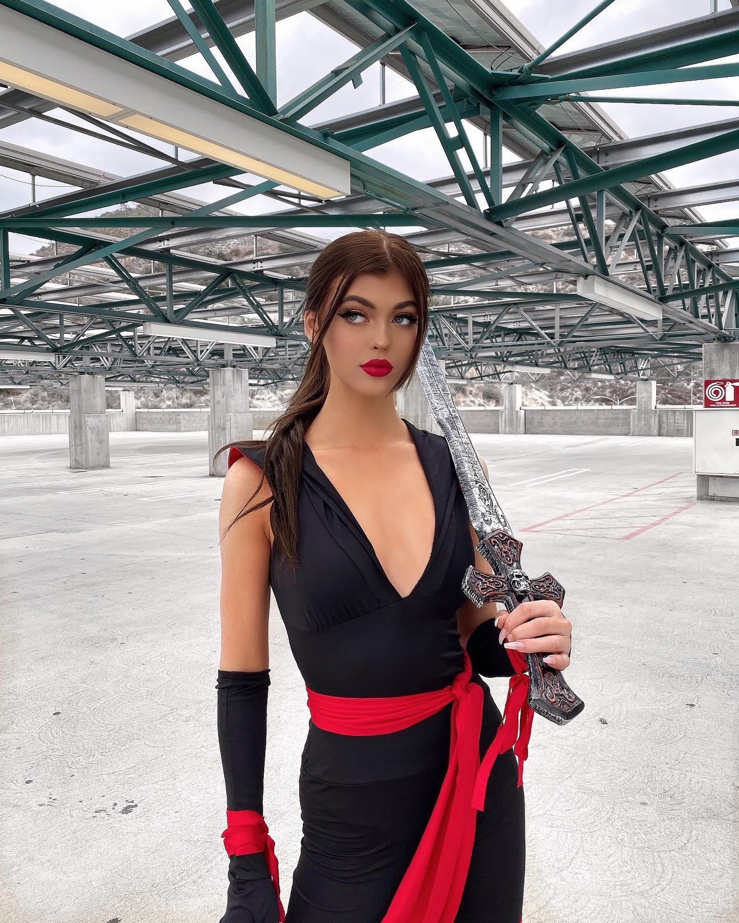 a woman dressed in a ninja costume stands in a parking garage holding a sword. she is wearing a black dress with a red belt, long gloves, and has her hair in a ponytail. she is also wearing red lipstick. the background is a parking garage with a metal ceiling and concrete pillars.