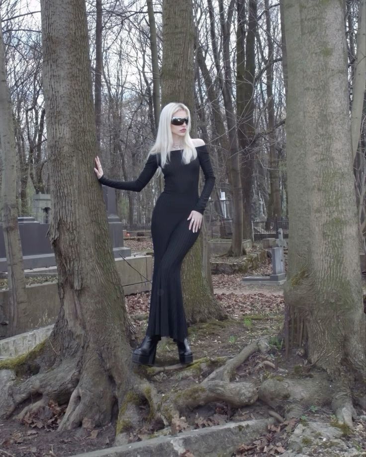 a blonde woman in a black offtheshoulder maxi dress and black platform boots poses in a cemetery.  she's wearing sunglasses and her style is gothic and mysterious. the setting is a wooded area with large trees and visible gravestones. the overall atmosphere is dark and moody.