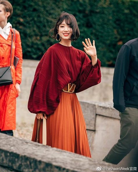 a young woman is walking down a cobbled street, waving at the camera. she is wearing a stylish red blouse and an orange skirt cinched at the waist with a gold buckled belt. she is also wearing large gold hoop earrings. the photo appears to have been taken during fashion week in paris.