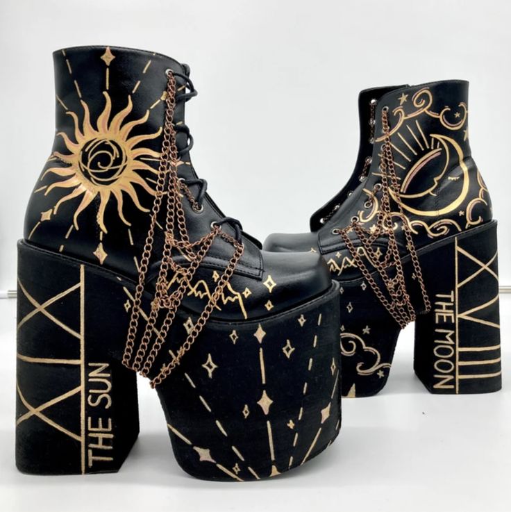 the image shows a pair of black platform boots adorned with gold tarot cardthemed designs. the left boot features a design of the sun with radiating lines, while the right boot displays a crescent moon with a sleeping face. each boot has the corresponding name of the tarot card, "the sun" and "the moon" respectively, painted on the heel. gold chains are draped across the laces and sides of the boots. the platforms of the boots also have decorative gold patterns with stars and other occult symbols.