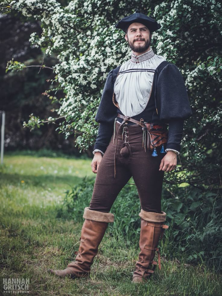 a man in a renaissance costume stands in a grassy field in front of a flowering bush. he is wearing a black hat, a doublet, hose, and boots. he also has a beard.