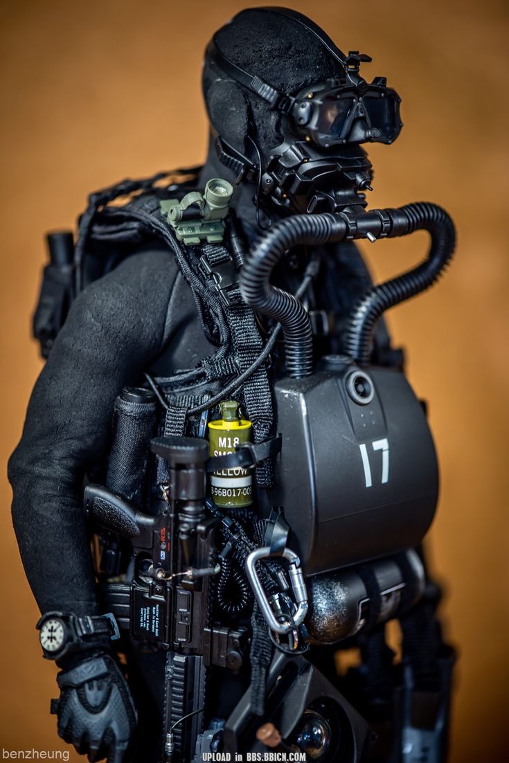 this is a closeup of an action figure dressed as a scuba diving soldier. he is wearing a black wetsuit, a black diving helmet with a black face mask, and a black tank on his back. he is also carrying a black rifle and other tactical gear. the action figure is standing in front of a brown background.
