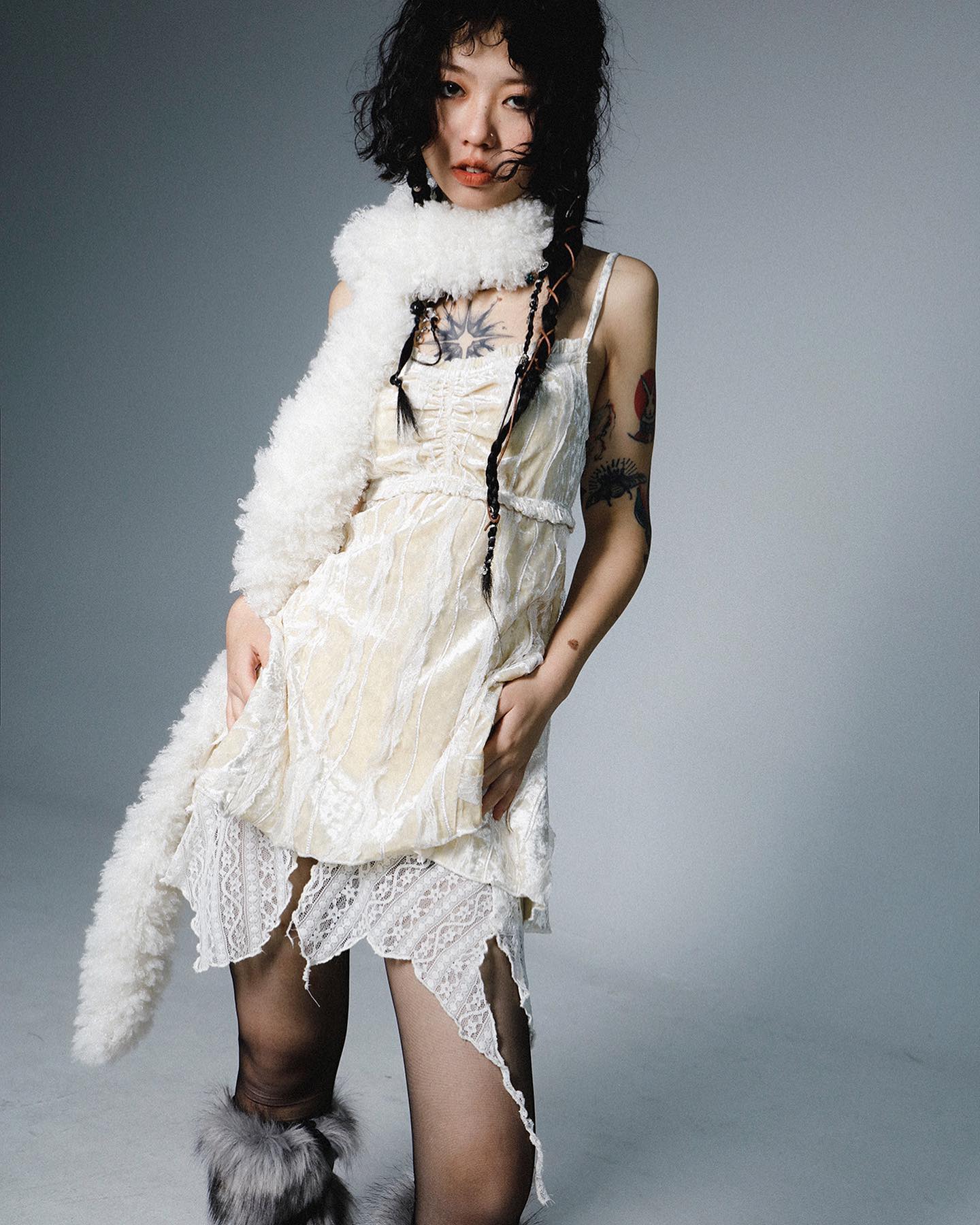 the image is a fashion portrait of a young woman with dark curly hair. she's wearing a stylish offwhite velvet dress with a lace overlay and a ripped hemline. the dress has a slightly bohemian or grunge aesthetic.  she's accessorized with a large, fluffy white scarf and furtrimmed boots. she has visible tattoos on her arms and chest. the overall style is edgy yet elegant, capturing a unique blend of textures and aesthetics. the background is a simple, neutral gray that allows the model and her outfit to stand out.  the lighting is soft and even, enhancing the details of the clothing and the model's features.