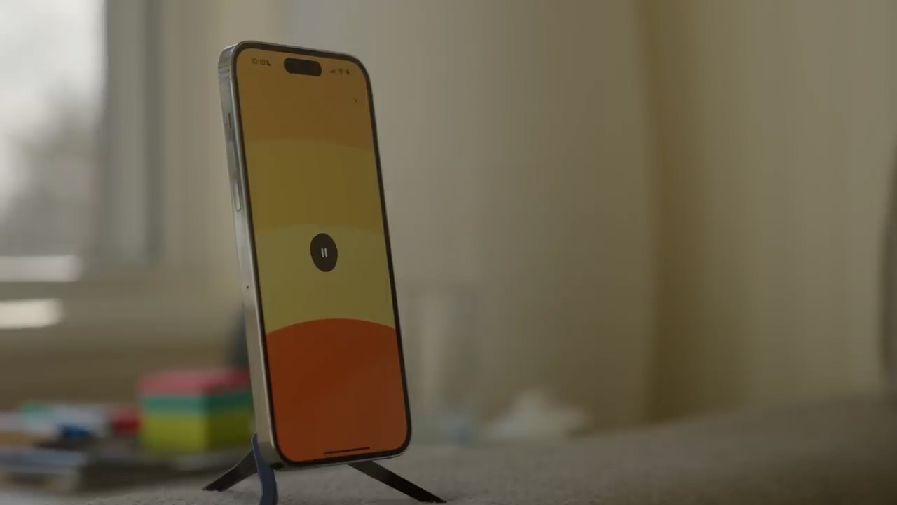 a silver iphone 14 pro is propped up on a black phone stand. the phone's screen is displaying a music player app. the background of the screen is an abstract gradient of yellow and orange. the room is brightly lit by natural light coming from a window on the left side of the image. the background is slightly out of focus, drawing attention to the phone in the foreground.