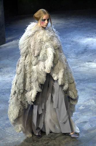 a fashion model is wearing a dramatic white and grey feather and fur dress. the model is standing on a runway and is looking down. the dress is long and flowing and has a cape attached to it. the model is wearing makeup and is looking down. the dress is very fluffy and looks like it is made of feathers.