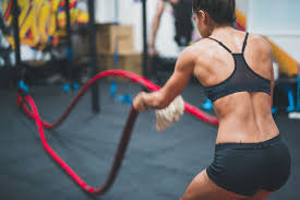 a woman is engaged in a fitness workout in a gym, using battle ropes. she is wearing a sports bra and shorts, revealing her muscular back and athletic build. the background shows other gym equipment and indistinct figures of people, suggesting a dynamic and active environment.