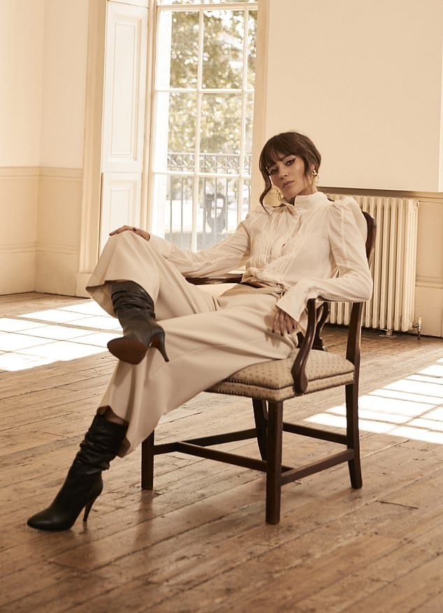 a fashion model is sitting on a chair in a room with a window. she is wearing a white shirt, beige pants, and black boots. her hair is styled in a chic way. she is looking at the camera. the room is decorated in a simple, elegant style. the image is high quality and has a professional look.