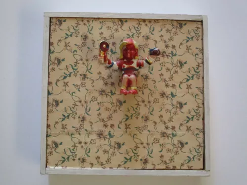 an art piece consisting of a square frame filled with a floral patterned background and a figurine attached in the center. the figurine, painted in red and white with yellow hair, holds a small object in each hand.