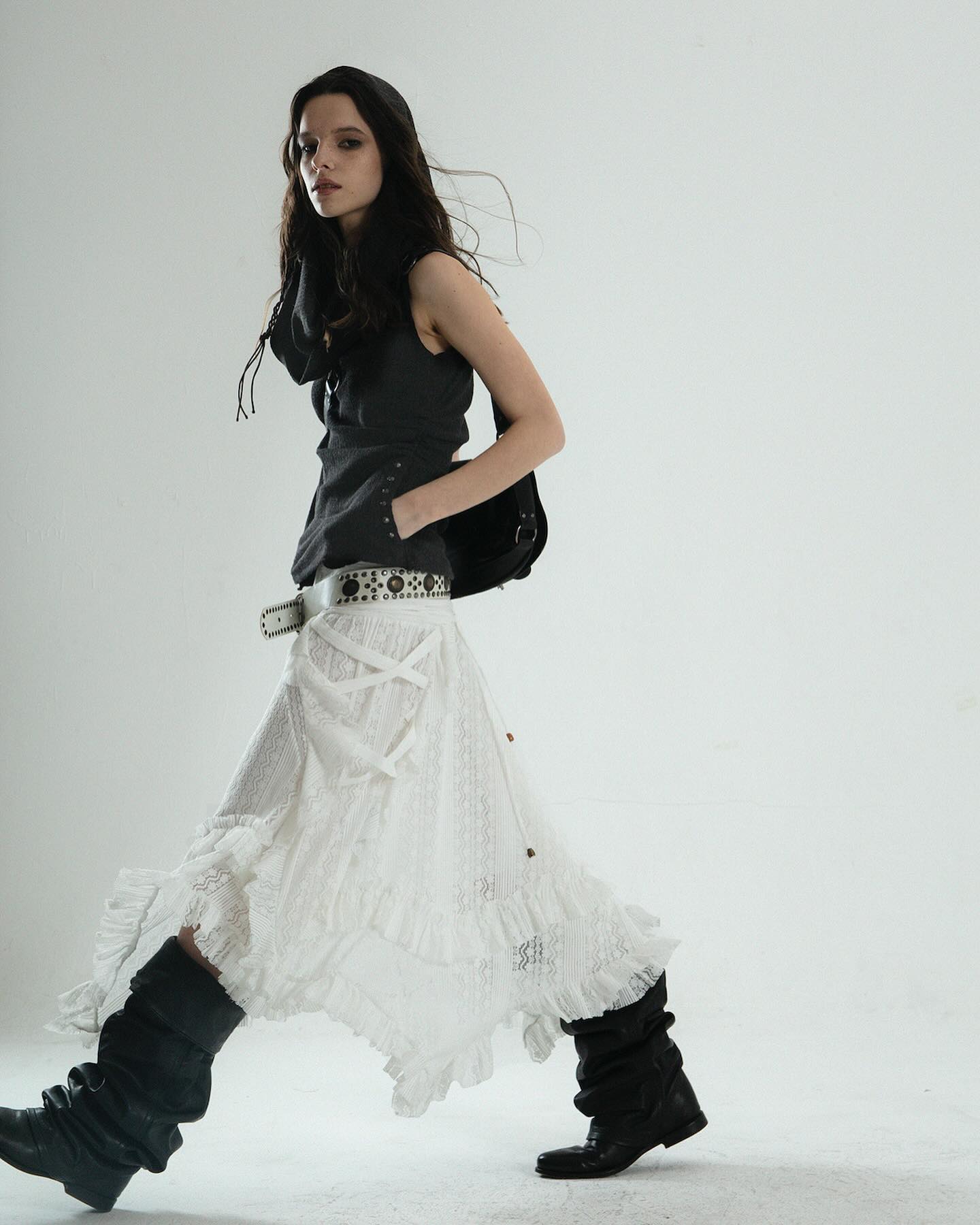 a fulllength studio shot of a female model showcasing a stylish outfit. she is wearing a white, asymmetrical, ruffled lace skirt with a black sleeveless hooded top.  the outfit is accessorized with a studded belt and black boots. the model is walking, her pose dynamic and expressive, against a plain white backdrop. the overall style leans towards a blend of gothic, bohemian, and alternative fashion influences.